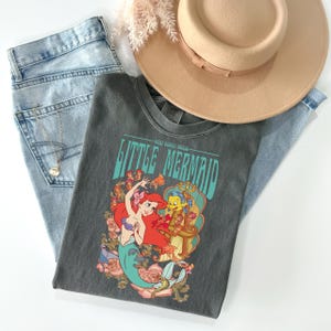 May include: A gray t-shirt featuring a colorful graphic of Ariel and Flounder from The Little Mermaid, with the text "Little Mermaid". The shirt is laid on top of light blue jeans, a tan hat, and a gold necklace.