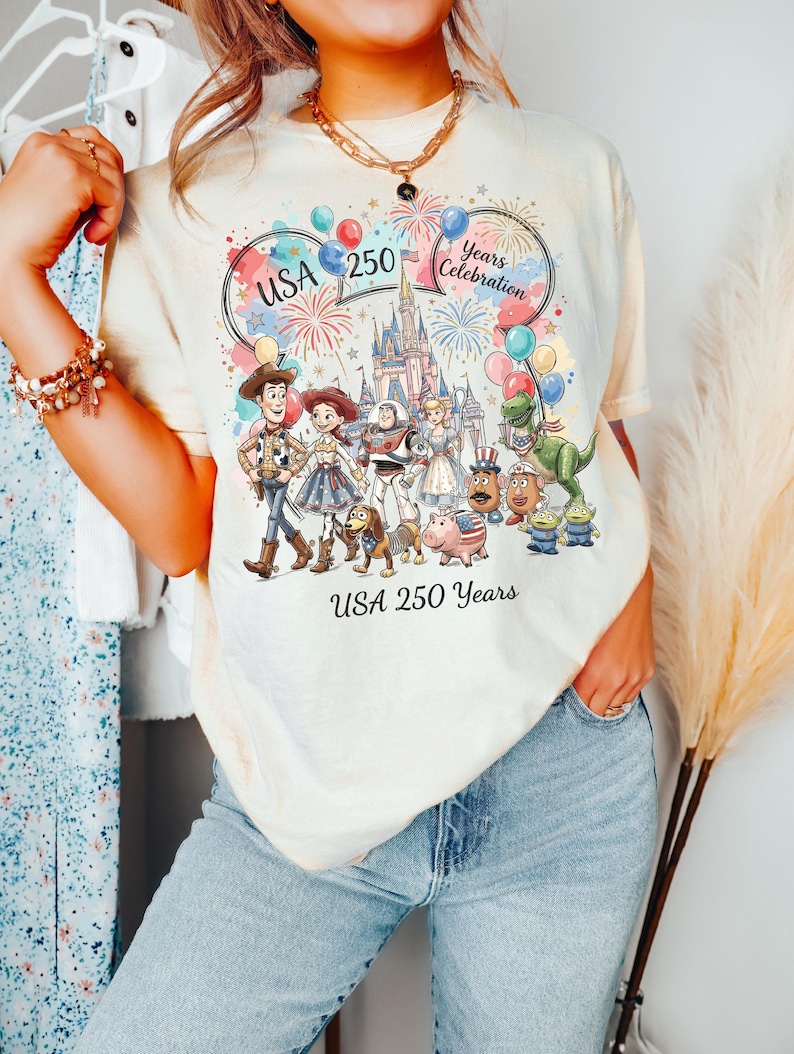 Peut inclure: Un t-shirt couleur cr&egrave;me avec un motif repr&eacute;sentant divers personnages anim&eacute;s, un ch&acirc;teau, des feux d'artifice et des ballons. Le texte indique USA 250 Years Celebration et USA 250 Years. Le design inclut Woody, Jessie, Buzz l'&Eacute;clair et autres.
