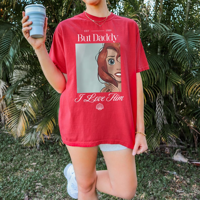 Puede incluir: Camiseta roja con el texto "But Daddy" y "I Love Him", con una ilustraci&oacute;n de dibujos animados. La camiseta tiene un estilo vintage. El dise&ntilde;o probablemente est&aacute; impreso en una camiseta de algod&oacute;n.