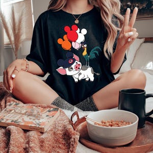 Pode incluir: T-shirt preto com um gr&aacute;fico colorido de personagens de desenhos animados, incluindo um porco com chap&eacute;u, um galo e bal&otilde;es vermelhos, laranja e brancos. A camisa tem um corte folgado, adequado para uso casual.