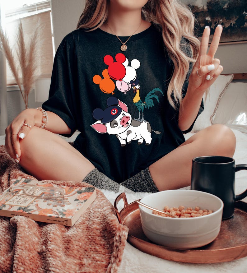 Pode incluir: T-shirt preto com um gr&aacute;fico colorido de personagens de desenhos animados, incluindo um porco com chap&eacute;u, um galo e bal&otilde;es vermelhos, laranja e brancos. A camisa tem um corte folgado, adequado para uso casual.