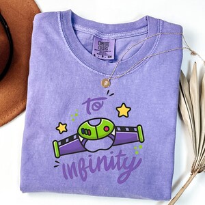May include: A lavender Comfort Colors t-shirt with a graphic of a cartoon spaceship and the words "to infinity" in a whimsical font. The design includes green, purple, and yellow accents. A gold necklace is draped across the shirt.