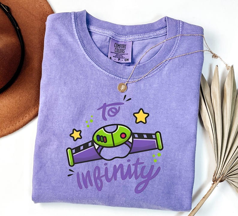 May include: A lavender Comfort Colors t-shirt with a graphic of a cartoon spaceship and the words "to infinity" in a whimsical font. The design includes green, purple, and yellow accents. A gold necklace is draped across the shirt.
