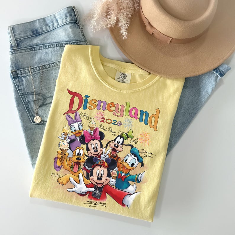 K&ouml;nnte beinhalten: Gelbes Disneyland 2026 Grafik-T-Shirt mit Mickey Mouse und Freunden. Das Shirt liegt auf hellblauen Jeans, einem beigen Hut und einer goldenen Halskette. Das T-Shirt hat einen Rundhalsausschnitt und kurze &Auml;rmel.