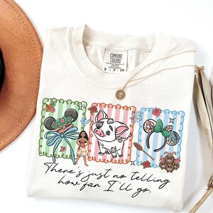 May include: A cream-colored t-shirt featuring a graphic with three vertical striped panels in green, pink, and blue. The design includes a character, a pig, mouse ears, and the text "There's just no telling how far I'll go." The shirt has a Comfort Colors label.