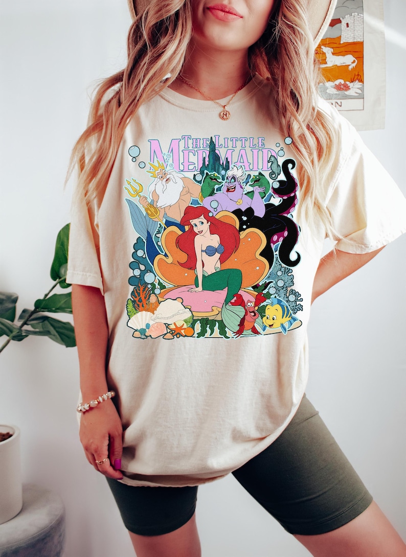 Op de afbeelding: Cr&egrave;mekleurig T-shirt met een kleurrijke afbeelding van personages uit "De Kleine Zeemeermin". Het ontwerp bevat Ariel, Koning Triton, Ursula en Botje. De woorden "The Little Mermaid" staan bovenaan in een gestileerd lettertype.