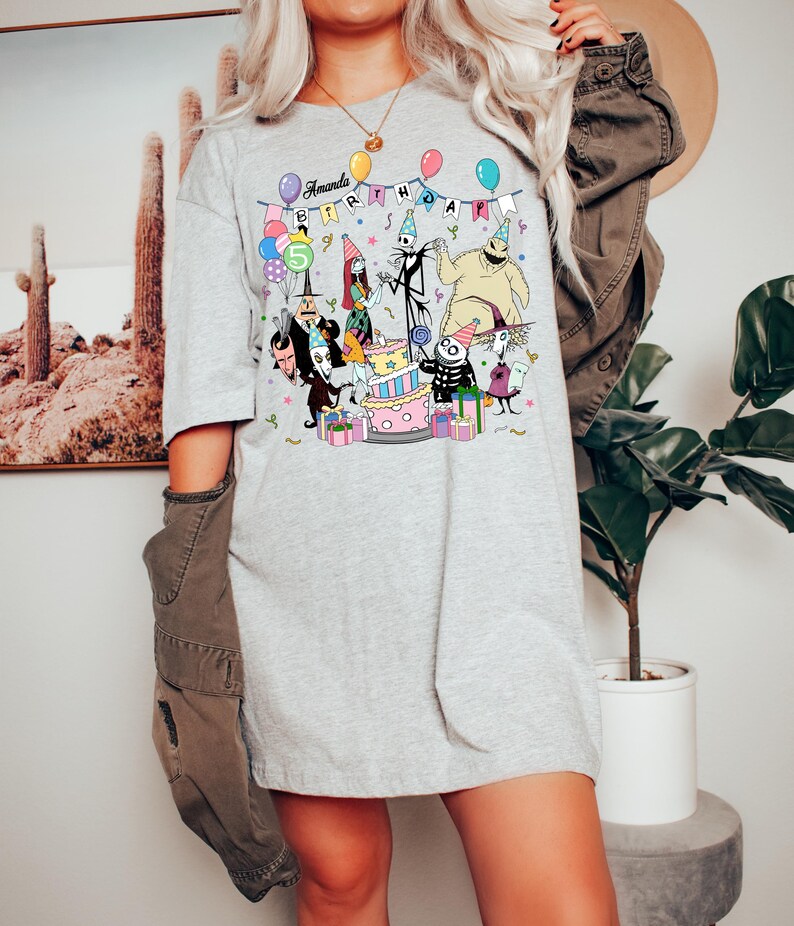 May include: A light grey t-shirt with a colourful graphic of characters from The Nightmare Before Christmas celebrating a birthday. The text "Amanda Birthday" is displayed above the characters, with a number "5" on a balloon.