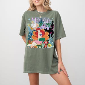 Op de afbeelding: Olijfgroen T-shirt met een kleurrijke afbeelding van personages uit "De Kleine Zeemeermin". Het ontwerp bevat Ariel, Koning Triton, Ursula en Botje. Het shirt heeft korte mouwen en een relaxte pasvorm.