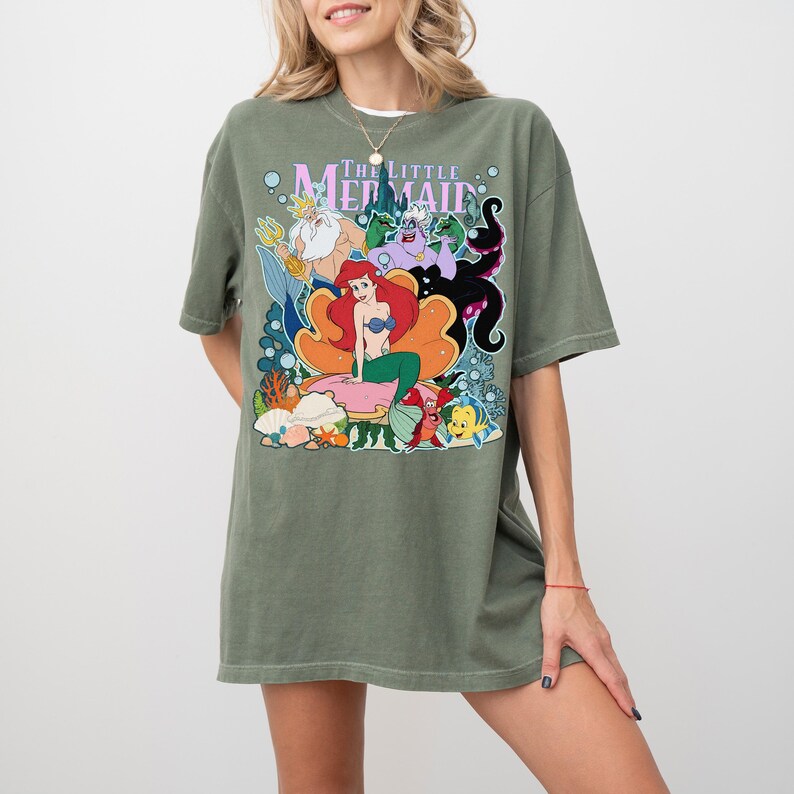 Op de afbeelding: Olijfgroen T-shirt met een kleurrijke afbeelding van personages uit "De Kleine Zeemeermin". Het ontwerp bevat Ariel, Koning Triton, Ursula en Botje. Het shirt heeft korte mouwen en een relaxte pasvorm.