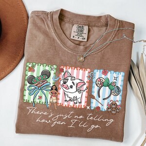 May include: A brown short-sleeve t-shirt featuring a graphic print with three rectangular panels containing a Moana-themed design, including a character, a pig, and ear headbands. The text "There's just no telling how far I'll go" is printed below.