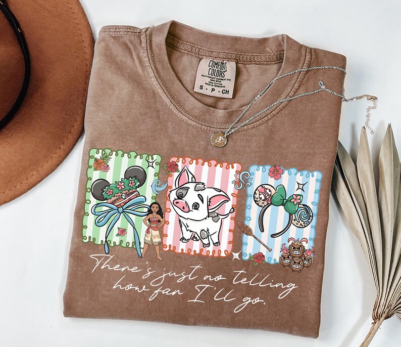 May include: A brown short-sleeve t-shirt featuring a graphic print with three rectangular panels containing a Moana-themed design, including a character, a pig, and ear headbands. The text "There's just no telling how far I'll go" is printed below.