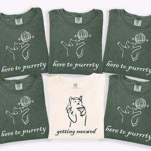 May include: A collection of green and cream-colored t-shirts. Most shirts feature a white graphic of a cat with a disco ball and the text "here to purrrty." One cream shirt has a cat with a martini and the text "getting meowed."