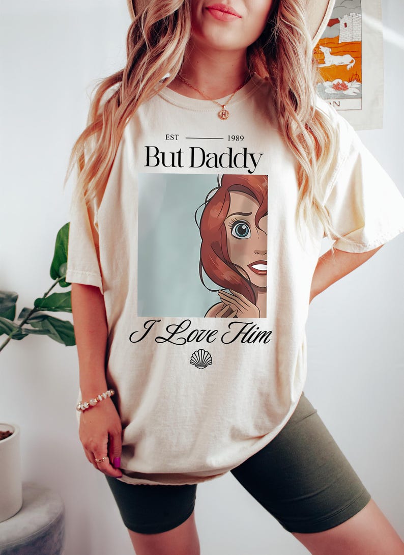 Puede incluir: Camiseta color crema con el texto "But Daddy I Love Him" y un dibujo animado. La camiseta tiene una est&eacute;tica vintage, con el a&ntilde;o "1989" impreso encima del texto. El dise&ntilde;o incluye un gr&aacute;fico de concha.