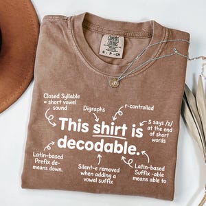 May include: A brown short-sleeved t-shirt with white text that reads: This shirt is decodable. The graphic includes arrows pointing to linguistic notes such as Closed Syllable, Digraphs, r-controlled, Latin-based Prefix de-, Silent-e, and Latin-based Suffix -able.