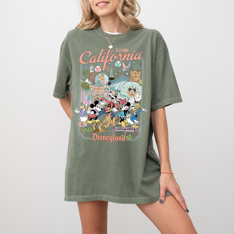 Puede incluir: Camiseta verde oliva con un dise&ntilde;o de Disneyland que presenta a Mickey Mouse y sus amigos. El dise&ntilde;o incluye el texto "California" y "Est. 1955" en un estilo retro. La camiseta es de corte holgado.