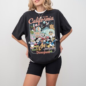 Puede incluir: Camiseta negra con un gr&aacute;fico colorido con personajes de Disneyland y el texto "California Disneyland Est. 1955". La camiseta tiene un aspecto vintage, ligeramente descolorido.