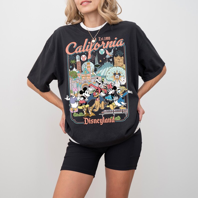 Puede incluir: Camiseta negra con un gr&aacute;fico colorido con personajes de Disneyland y el texto "California Disneyland Est. 1955". La camiseta tiene un aspecto vintage, ligeramente descolorido.