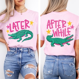 Peut inclure: T-shirt rose avec un motif d'alligator vert et le texte "LATER" à l'avant et "AFTER WHILE" à l'arrière. Le design comprend des étoiles jaunes. Le t-shirt est porté avec un jean bleu.
