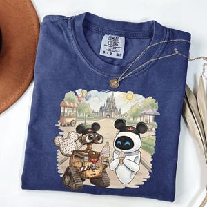 Puede incluir: Una camiseta azul marino Comfort Colors con un gr&aacute;fico colorido de Wall-E y Eva de la pel&iacute;cula de Disney, disfrutando de un helado frente a un castillo. La camiseta tiene una peque&ntilde;a etiqueta en el cuello y un collar dorado.