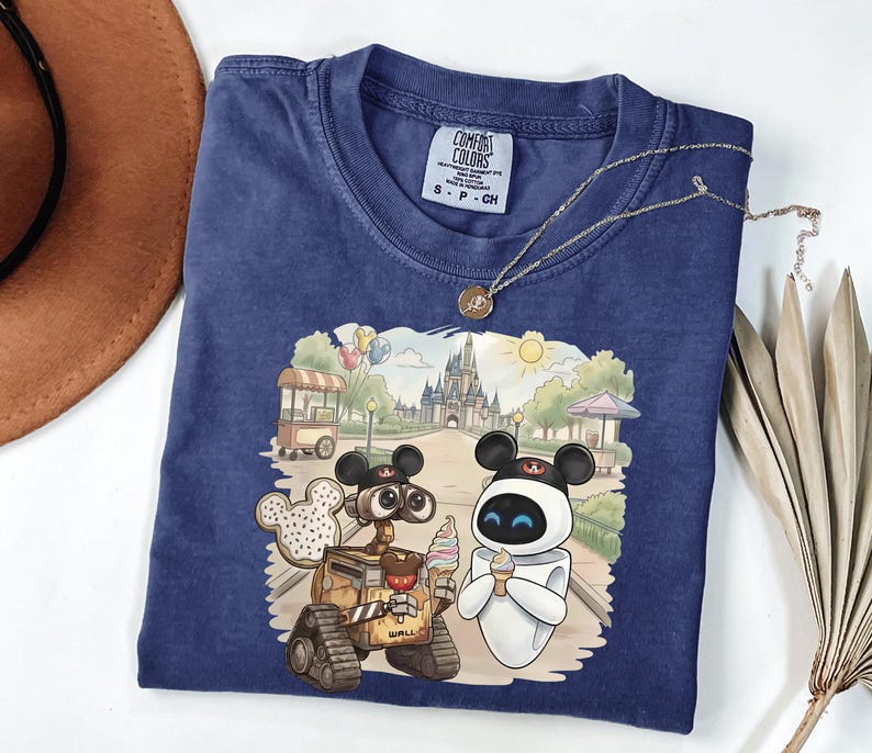 Puede incluir: Una camiseta azul marino Comfort Colors con un gr&aacute;fico colorido de Wall-E y Eva de la pel&iacute;cula de Disney, disfrutando de un helado frente a un castillo. La camiseta tiene una peque&ntilde;a etiqueta en el cuello y un collar dorado.