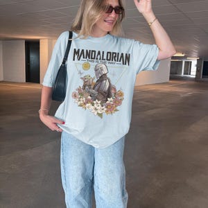 May include: A light blue t-shirt featuring a graphic print of The Mandalorian and Grogu surrounded by colorful flowers and butterflies. The text "MANDALORIAN" and "THIS IS THE WAY" is printed in black above the characters within a thin gold triangle.