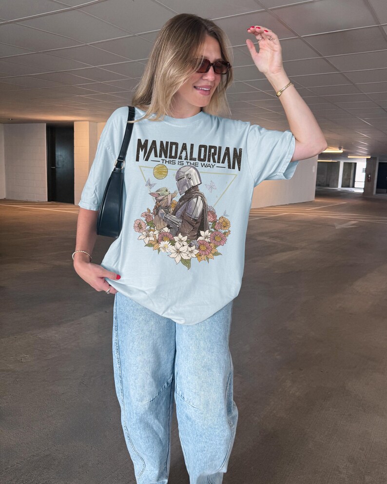 May include: A light blue t-shirt featuring a graphic print of The Mandalorian and Grogu surrounded by colorful flowers and butterflies. The text "MANDALORIAN" and "THIS IS THE WAY" is printed in black above the characters within a thin gold triangle.