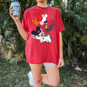 Pode incluir: Camiseta vermelha com uma ilustra&ccedil;&atilde;o de desenho animado de um porco usando um chap&eacute;u, um galo e bal&otilde;es em forma de Mickey Mouse. O porco tem manchas pretas e brancas. O galo &eacute; verde, amarelo e azul. Os bal&otilde;es s&atilde;o vermelhos, laranja e brancos.