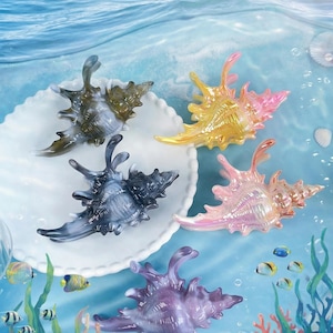 Op de afbeelding: Een assortiment kleurrijke, doorschijnende schelpenbeeldjes in tinten grijs, geel, roze en paars. De schelpen zijn gerangschikt op een wit, geschulpte bord, met een onderwaterachtergrond met koraal en vissen.