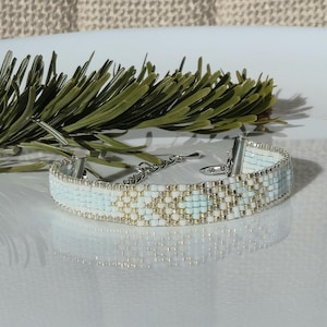 Könnte beinhalten: Ein Armband mit Perlen und einem silbernen Verschluss. Das Armband hat ein Muster aus hellblauen, weißen und goldenen Perlen. Das Armband ist etwa 2 cm breit. Ein Zweig mit grünem Laub befindet sich im Hintergrund.