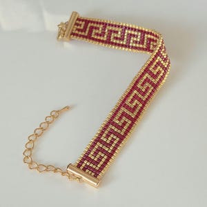 Könnte beinhalten: Ein Armband aus Perlen in Burgunderrot und Gold mit einem geometrischen Muster. Das Armband hat eine goldfarbene Kette und einen Verschluss zur Größenanpassung. Das Design ist ein sich wiederholendes griechisches Schlüsselmuster.