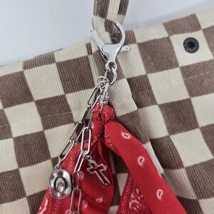 May include: A corduroy tote bag with a brown and cream checkerboard pattern. A silver chain with charms, including a cowboy hat, cross, and sunflower, hangs from the bag's handle. A red bandana with a white paisley design is attached.