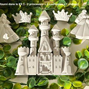 Peut inclure: Un château blanc, deux figurines de princesses (6 cm de haut) et deux couronnes (3,5 cm de large) sont présentés sur un lit de feuilles vertes. Le château mesure 15 cm x 12 cm. Le texte indique "Produit fourni dans le Kit 3 - 2 princesses + 2 couronnes + 1 château."