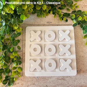 Peut inclure: Un jeu de tic-tac-toe blanc avec les lettres X et O. Le plateau mesure 21 cm de côté. Le texte "Produit fourni dans le Kit 1 - Classique" est en haut. Du feuillage vert est en arrière-plan.