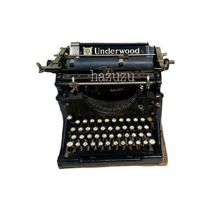 Puede incluir: Una máquina de escribir Underwood vintage negra con un diseño de teclado estándar. La máquina de escribir tiene un marco de metal y una palanca de retorno de carro negra.