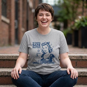 Puede incluir: Una camiseta gris con un diseño retro que presenta a una mujer sosteniendo una taza de café. El texto en la camiseta dice "Of course size matters" y "No one wants a small cup of coffee."