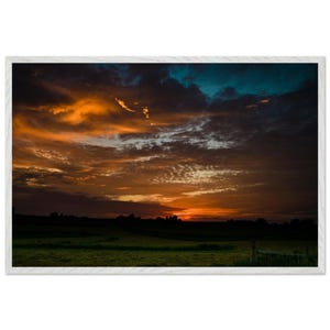 May include: A framed landscape photograph featuring a dramatic sunset with vibrant orange and deep blue clouds stretching across the sky above a dark, silhouetted field. The scene is enclosed within a simple, light-colored wooden frame.
