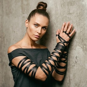 Op de afbeelding: Een persoon draagt een zwarte off-the-shoulder top en zwarte arm wraps. De arm wraps zijn gemaakt van leer en zijn gekruist rond de armen en vingers. De persoon heeft haar haar in een knot en leunt tegen een grijze muur.