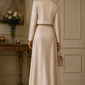 May include: A two-piece set in a light beige colour consisting of a cropped blazer with long sleeves and a matching floor-length maxi skirt. The outfit is paired with a small, structured handbag in a coordinating beige tone and high-heeled shoes.