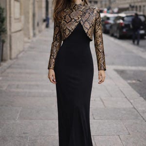 Puede incluir: Una mujer con un vestido largo negro y una chaqueta bolero con estampado de piel de serpiente. La chaqueta es marr&oacute;n y beige con cuello alto. El vestido es largo y ajustado. El fondo es una calle de la ciudad.