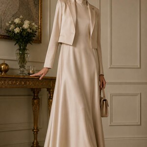 May include: A champagne-coloured satin ensemble consisting of a floor-length sleeveless dress with a high neckline and a matching cropped long-sleeve blazer with a notched lapel. The outfit is paired with a small structured handbag and pointed heels.