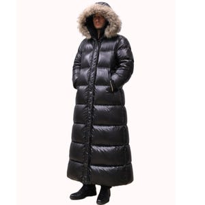 May include: A long, black puffer coat with a fur-trimmed hood. The coat extends to the ankles and features a full-length zipper. The person is wearing black boots. The coat is designed for cold weather.