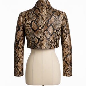 Puede incluir: Chaqueta corta con estampado de piel de serpiente en tonos marr&oacute;n y negro. La chaqueta, con mangas largas y cuello, se exhibe en un maniqu&iacute;. El dise&ntilde;o de la chaqueta imita la apariencia de las escamas de serpiente.