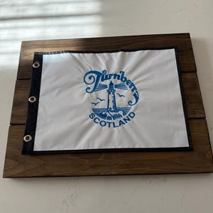 Puede incluir: Una bandera blanca con las palabras "Turnberry Scotland" en azul, con un faro y pájaros, montada sobre un marco de madera teñida oscura. La bandera tiene un borde negro y ojales metálicos para colgar. El marco de madera es rectangular.
