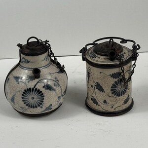 Può includere: Due antichi contenitori in ceramica con dettagli in metallo scuro. I contenitori sono bianchi con un motivo floreale blu. Uno ha una forma bulbosa, l'altro è cilindrico. Entrambi hanno coperchi e catene.