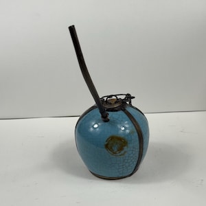 Può includere: Un recipiente rotondo in ceramica blu chiaro con un lungo beccuccio curvo marrone scuro. Il recipiente ha una glassa craquelé ed è accentuato da fasce metalliche scure e una parte superiore in metallo. Il design generale ricorda una lampada a olio antica.
