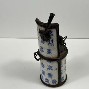 Può includere: Un antico contenitore cilindrico in ceramica con struttura in metallo. La ceramica bianca presenta un motivo floreale blu e caratteri cinesi. Il contenitore ha un coperchio incernierato e un manico. Il design ricorda una piccola stufa o lanterna.