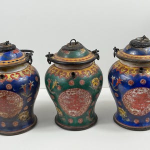 Può includere: Tre vasi antichi in ceramica con coperchi. I vasi sono dipinti con disegni intricati in blu, verde e giallo, con accenti rossi. Ogni vaso ha un manico e un coperchio in metallo e un motivo decorativo sul davanti.