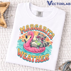 May include: White Comfort Colors t-shirt with a colorful graphic. The graphic features a raccoon wearing sunglasses and a flower crown, floating on a pink flamingo floatie, holding a margarita. The text "Margarita Weather" is arched above and below the image.