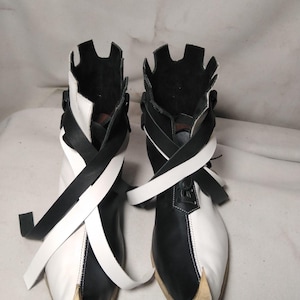 May include: A pair of ankle boots featuring a split black and white color-blocked design. The boots have a pointed toe, a notched collar, and long, flat wrap-around straps in contrasting black and white colors. The soles are light-colored.