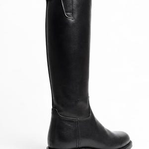 Könnte beinhalten: Ein schwarzer kniehoher Stiefel mit glatter Lederoptik, abgerundeter Schuhspitze, flachem Absatz und einer vertikalen Naht am hinteren Schaft. Am oberen hinteren Ende des Stiefels befindet sich ein kleiner Schnallenverschluss aus Metall.
